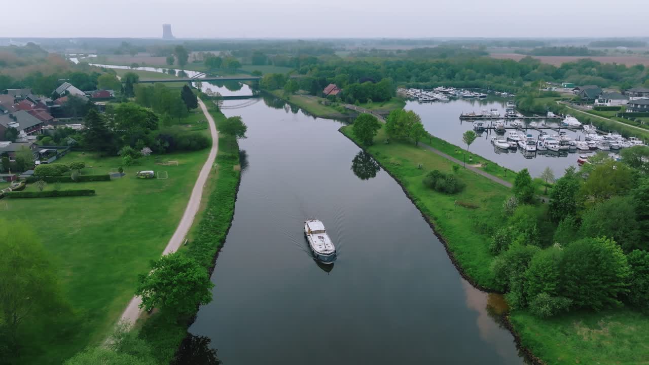 Zoom out revealing a barge sailing between green canal banks near a marina in northern Germany. Calm weather and gentle water reflections.