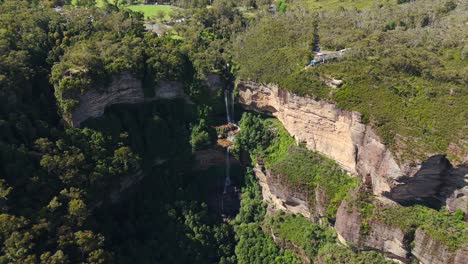 aerial left orbit reveals katoomba falls cascading down sandstone cliffs into lush forest, set within australia’s scenic blue mountains national park.