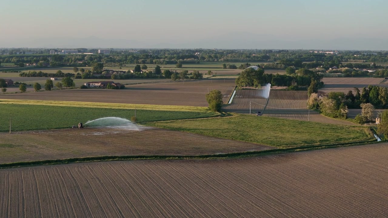 Irrigatori innaffiano terreni agricoli coltivati nella Pianura Padana vicino a Cremona, Lombardia, Italia, sorvolo lento in avanti con drone sopra campi agricoli e un paesaggio rurale di pianura durante la luce serale.
