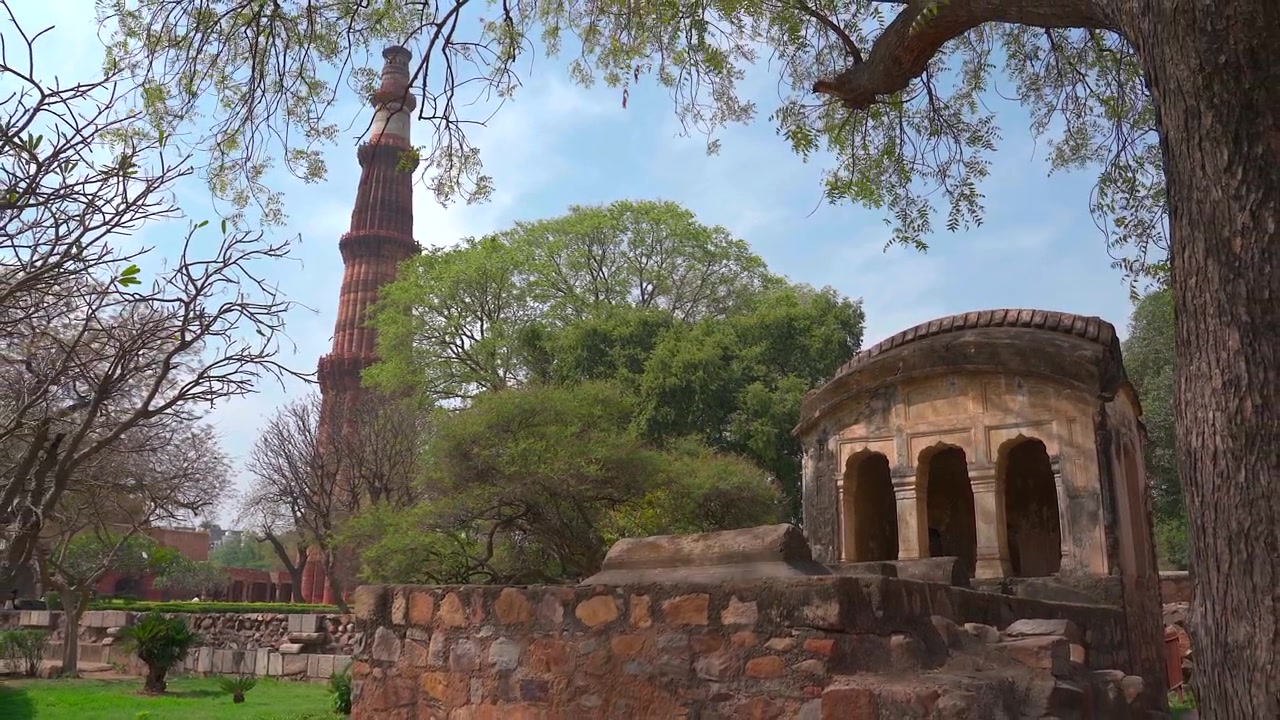 Inquadratura ampia statica dell'iconico minareto Qutub Minar incorniciato da alberi verdi e un antico padiglione in pietra nel complesso di Qutb a Nuova Delhi, India