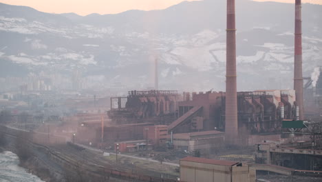 the fading light of dusk sets over an industrial steelworks, with towering chimneys and a snow-capped mountainous horizon