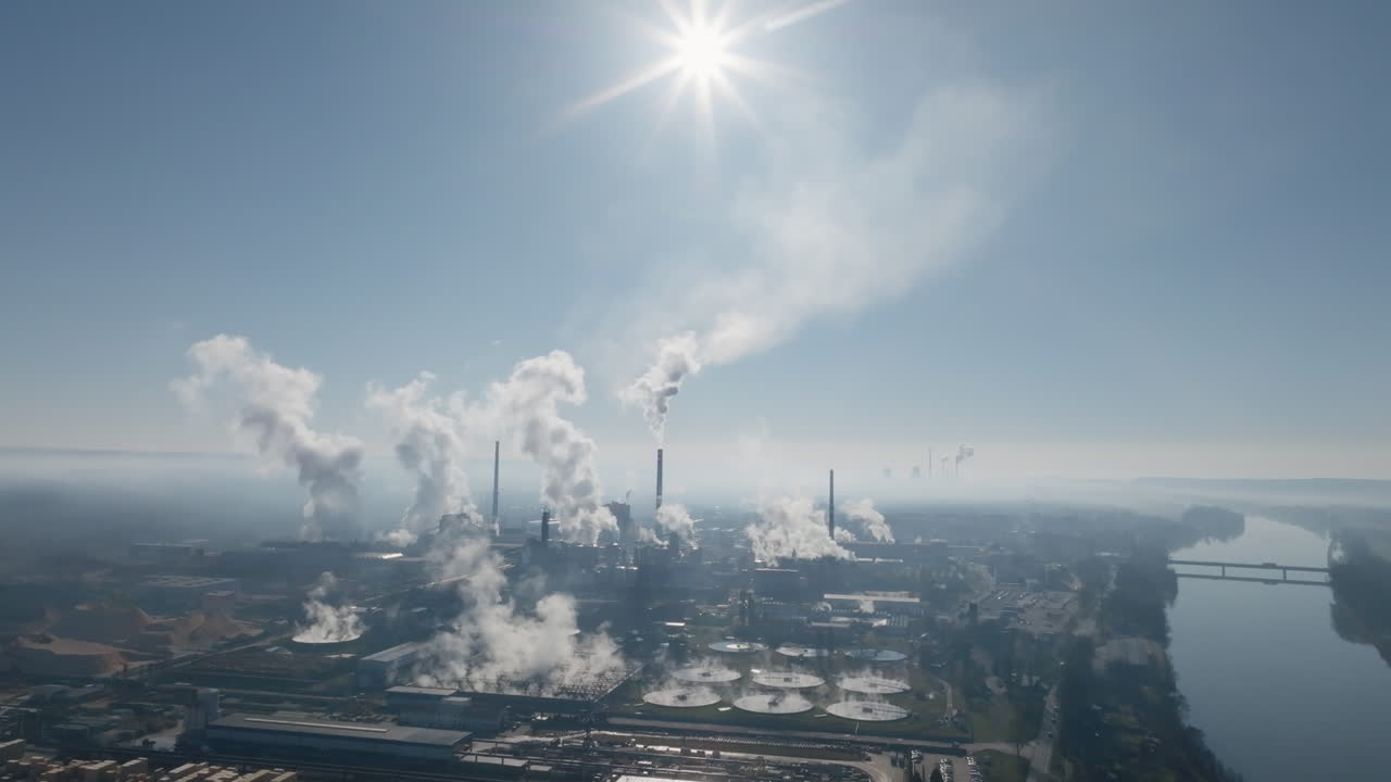 Veduta aerea da drone che mostra più camini che rilasciano vapore sopra un vasto complesso industriale, con bacini industriali e tetti visibili sotto la luce diurna