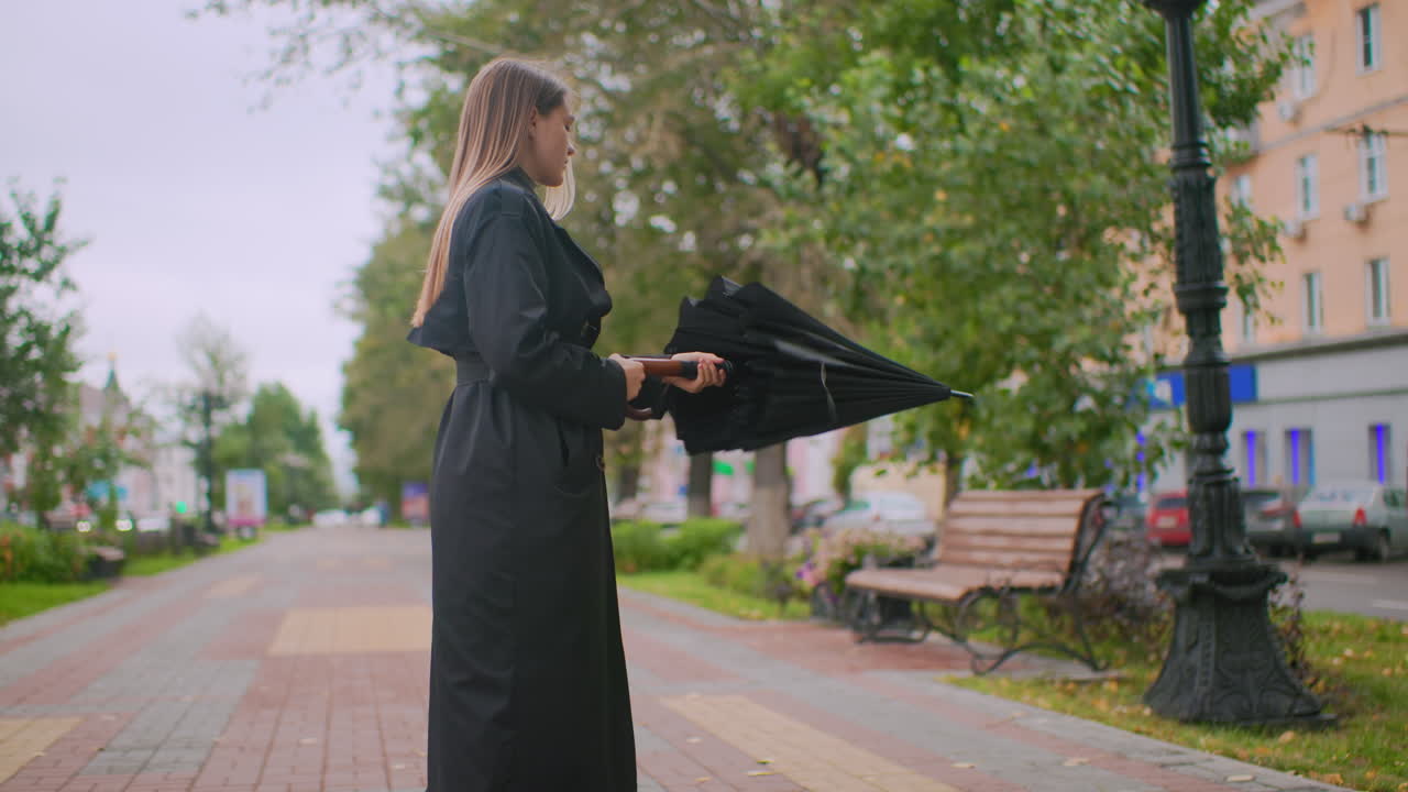 Pretty lady in long black coat hold and turn closed umbrella standing on city sidewalk near bench and lamppost in overcast weather with trees and buildings in background urban lifestyle
