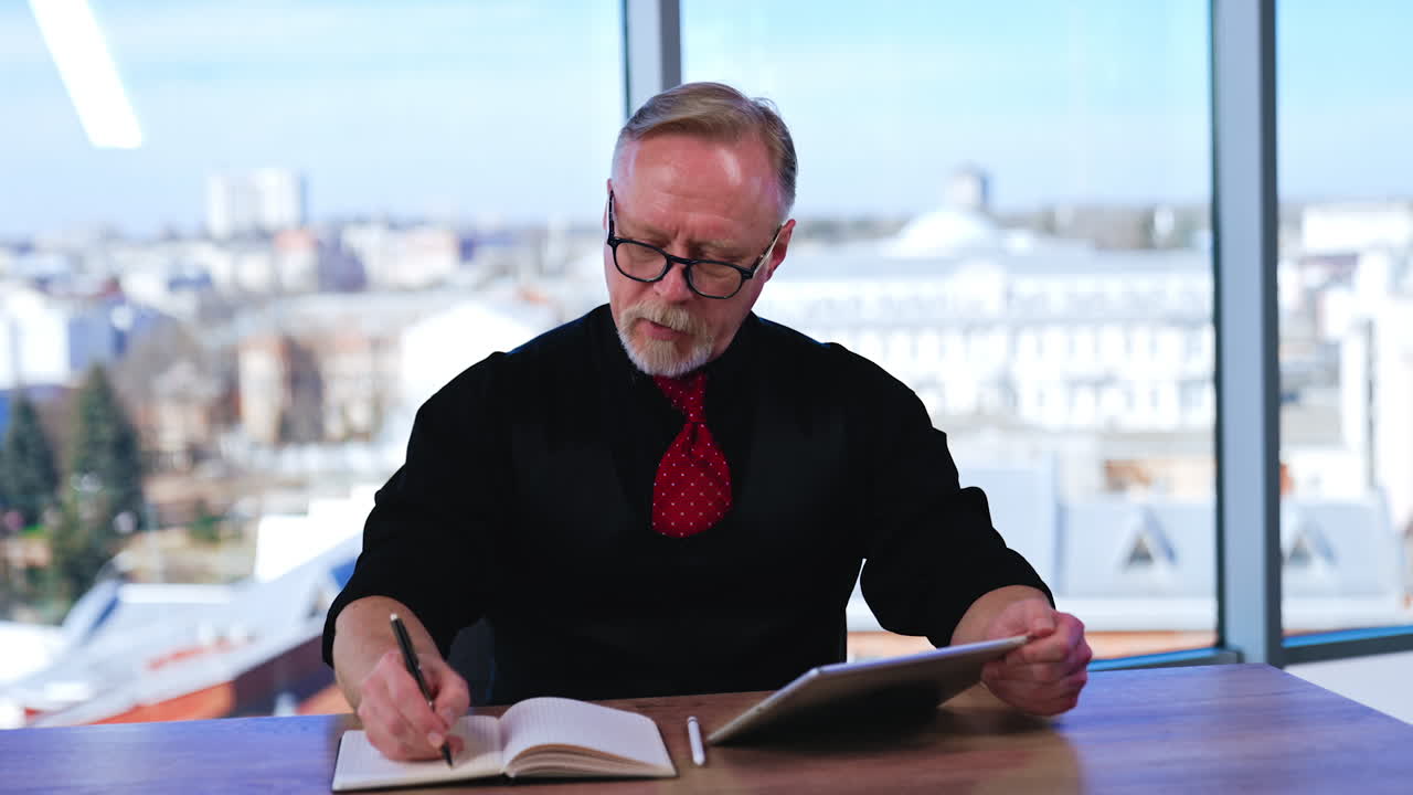 Businessman in black shirt, vest and red tie sits at desk writing in paper book. Man uses I-pad looking for data. Blurred window at backdrop.