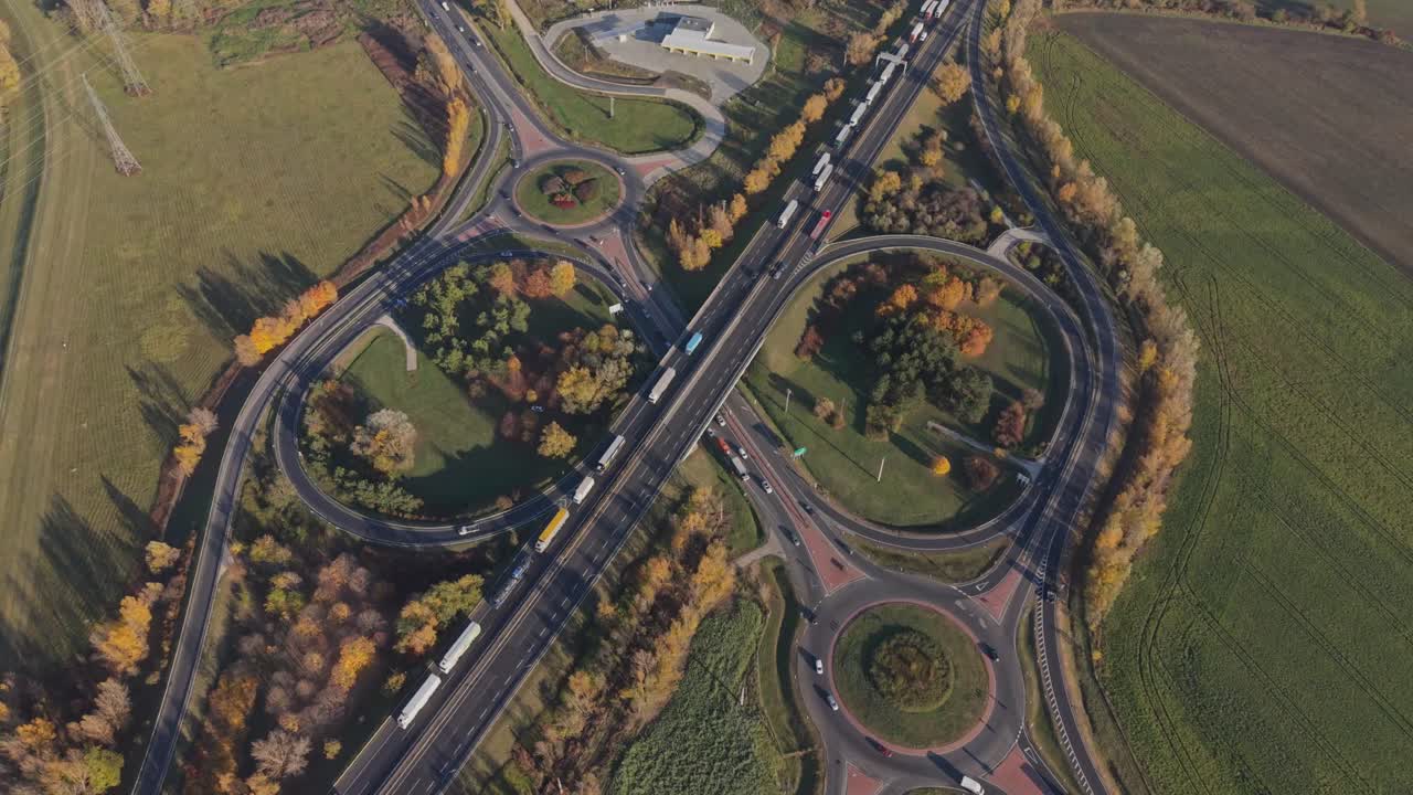 A high-angle aerial view of a large highway interchange with multiple roundabouts and overpasses. Trucks and cars travel through the junction surrounded by green fields and autumn-colored trees