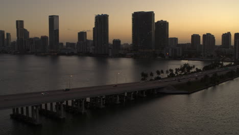 Miami-Bridge-Mit-Vorbeifahrenden-Autos-Und-Der-Innenstadt-Von-Miami-Im-Hintergrund-Bei-Dämmerung.-Luftaufnahme