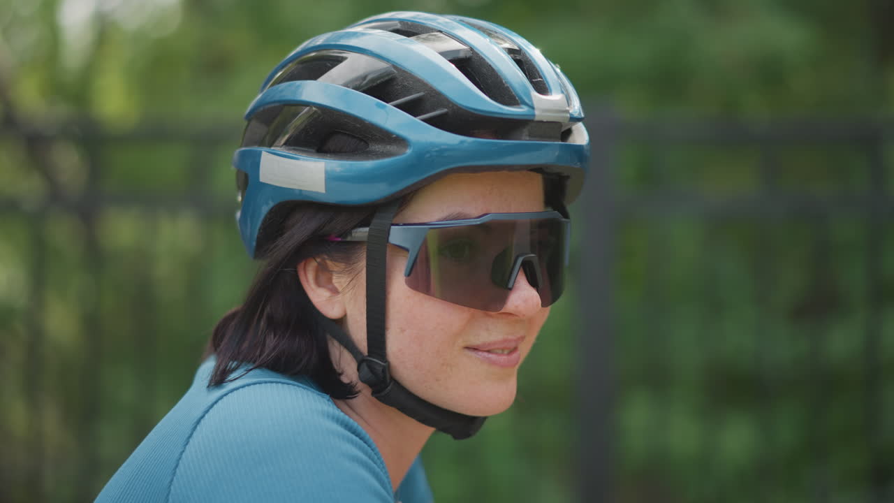 woman cyclist in blue helmet profile, close up side view of woman wearing sunglasses riding through green park, calm expression with subtle head turns, training and commuter mood, outdoor fitness