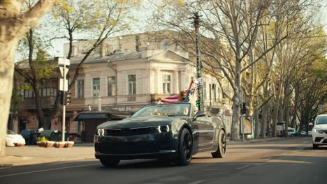 Ein-Dunkelgraues,-Glänzendes-Cabrio-Fährt-Eine-Sonnige-Straße-Entlang.-Ein-Mädchen-Sitzt-In-Einem-Cabrio-Und-Schwenkt-Die-Flagge-Der-Vereinigten-Staaten-Von-Amerika