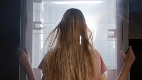 a young woman looks into an open refrigerator, where light shines and fog comes from. extreme cooling and coolness