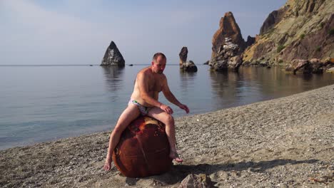 man doing exercise outdoors near to an old rusty floating marine mine on the beach with rocky shore and sea background. healthy lifestyle, pollution, nature protection, war and peace concept