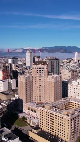 Vertical-Aerial-View,-San-Francisco-USA,-American-Flag-Waving-on-Downtown-Building-With-Golden-Gate-Bridge-and-Bay-in-Background