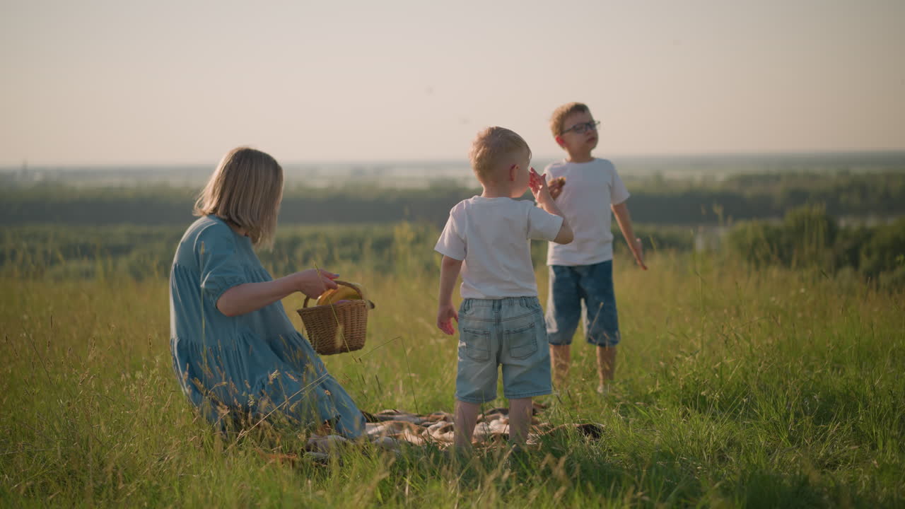 A mother in a blue dress bends down ,takes a basket of fruit while her two young sons, both in white tops, stand nearby in a grassy field
