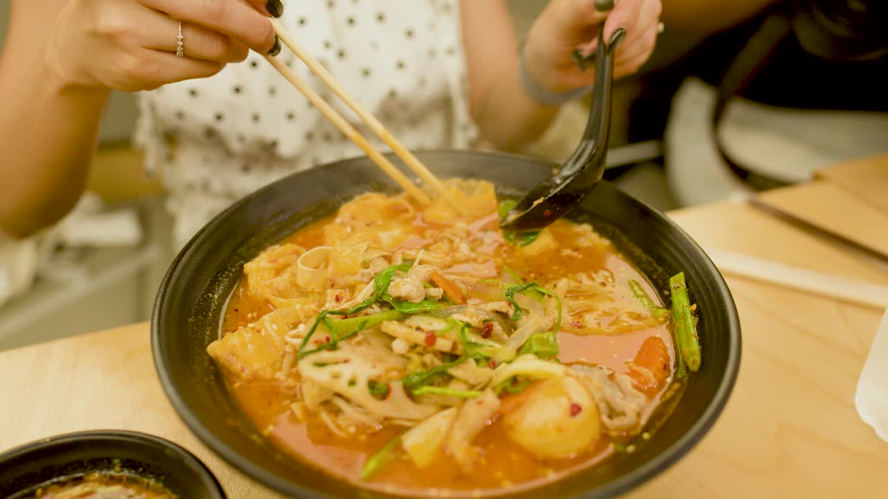 Close-up of woman using chopsticks and spoon to lift spicy noodle soup in warm lighting