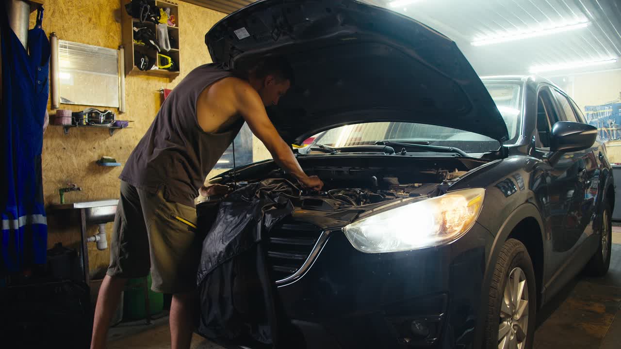 un mecánico maestro con un corte de pelo corto en una camiseta gris y pantalones cortos verdes repara el capó de un coche con los faros encendidos y una ligera bruma en su taller de garaje