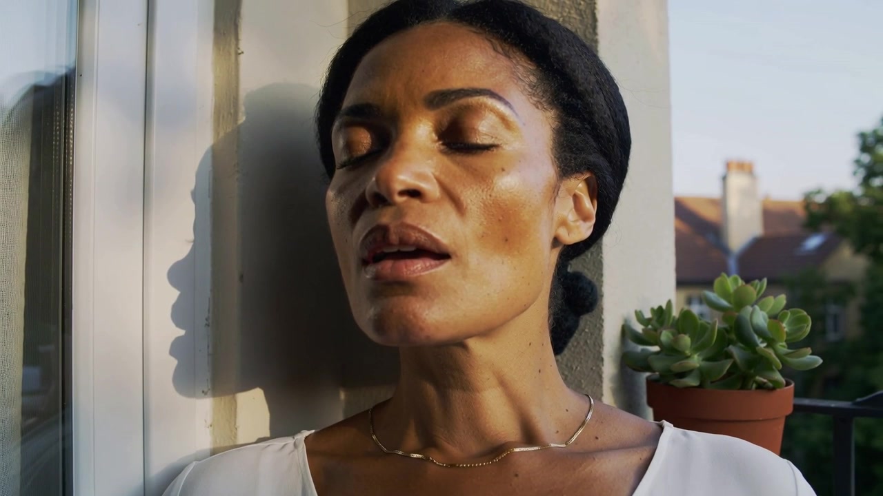 Breathing woman in light top and necklace leaning on doorframe balcony, succulent, sunlight calming