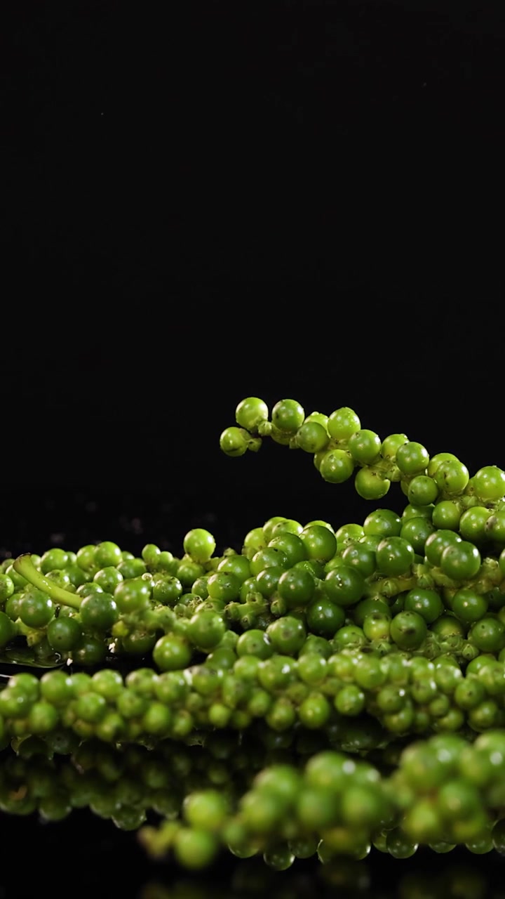 Grappoli di pepe verde fresco riposano su una superficie riflettente mentre goccioline d'acqua cadono al rallentatore, illuminate da un'illuminazione drammatica da studio su uno sfondo nero.