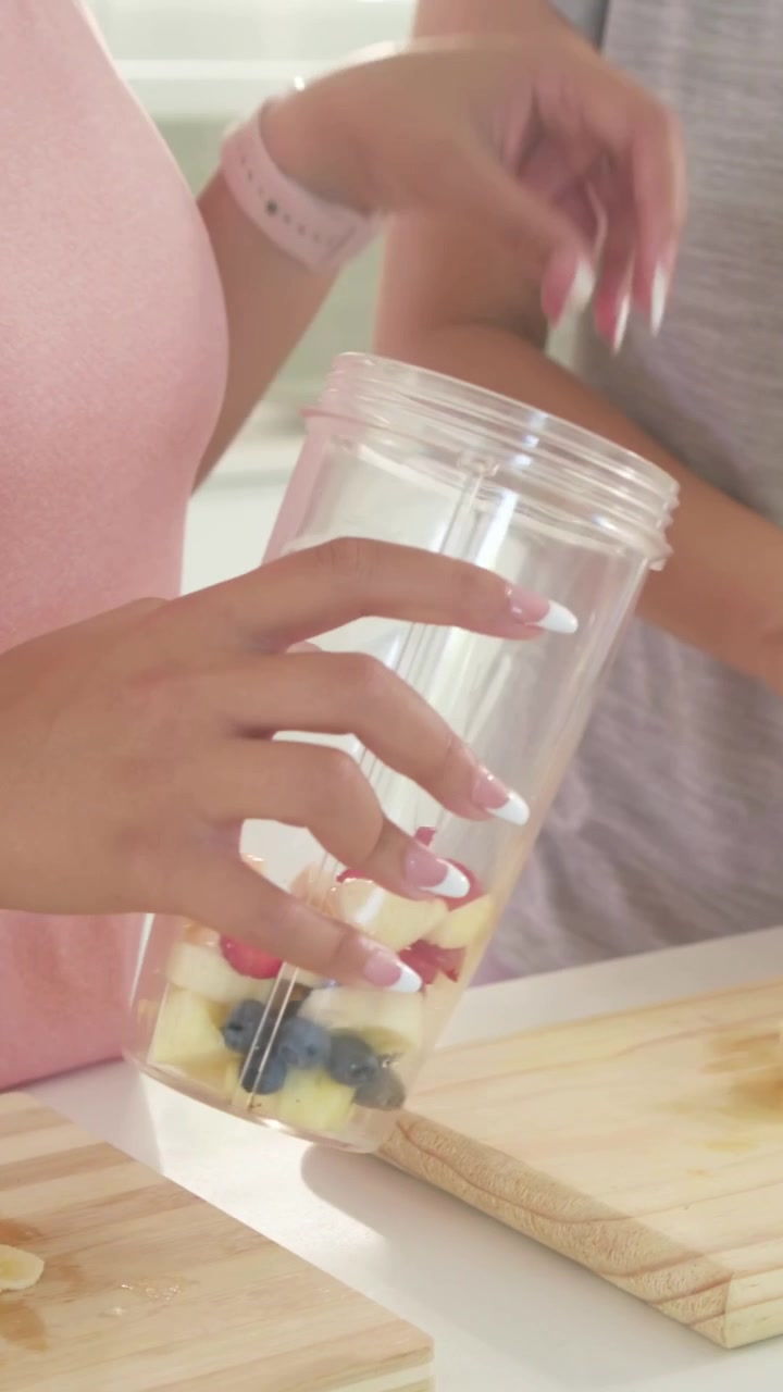 Vídeo vertical: Mulher de blusa rosa vendo o copo do liquidificador, pegando fruta enquanto amiga pica em casa para bater.