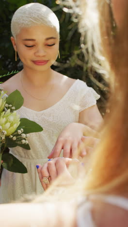vertical video of happy diverse female couple with bunch of flowers wearing ring in garden