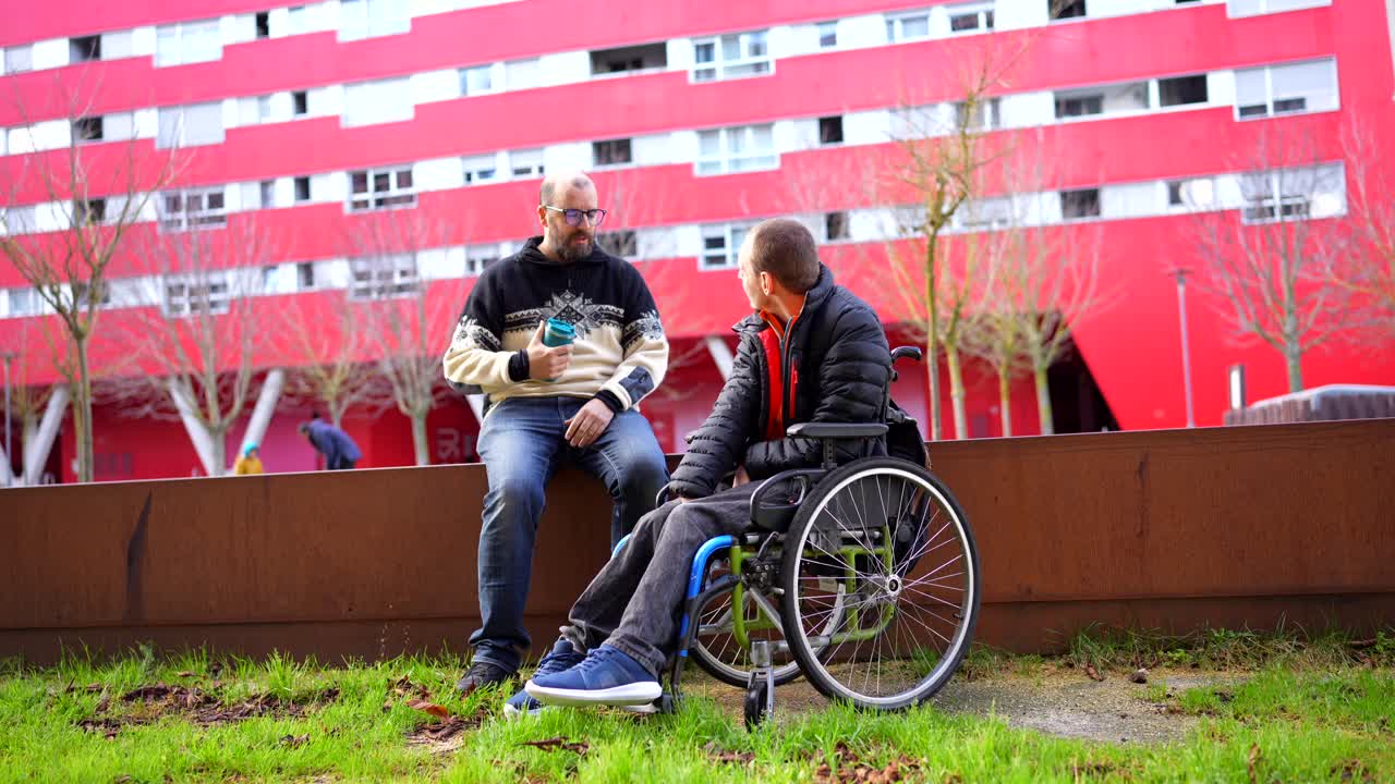 Two men, one in a wheelchair, sitting and talking outdoors in front of a red building