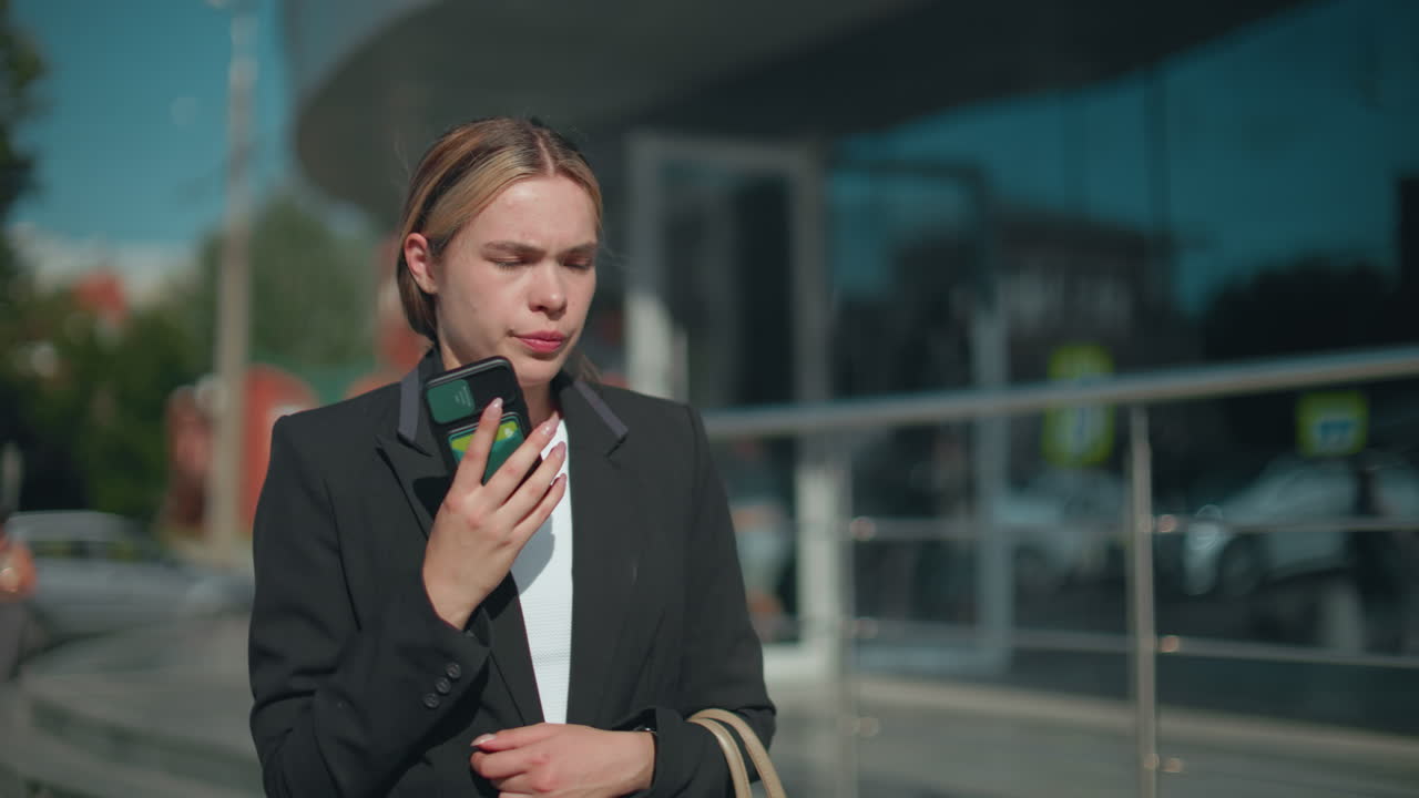 Upset woman in professional attire stands outdoors holding phone, appearing frustrated during call in sunny urban setting, blur background features mall entrance with passerby and modern railing