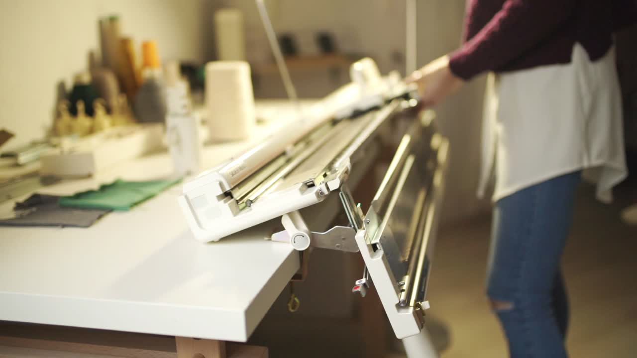 Jovem mulher ajusta máquina de tricô para trabalhar na oficina em casa.