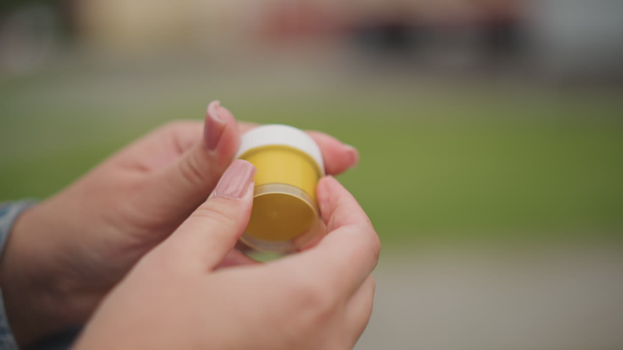 Outdoor closeup hands opening yellow jar, careful inspection of small plastic container on park bench, gentle fingernails twisting white lid, studying label and dosage, serene wellness routine
