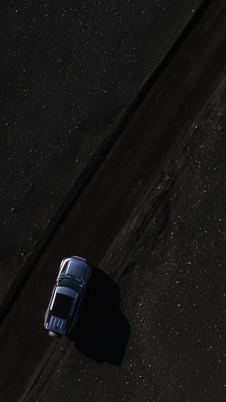 Toma cenital con dron de un SUV que empieza a conducir por una pista de arena volcánica negra, dejando marcas de neumáticos sobre un paisaje oscuro texturizado. Escena de viaje off-road minimalista y cinematográfica