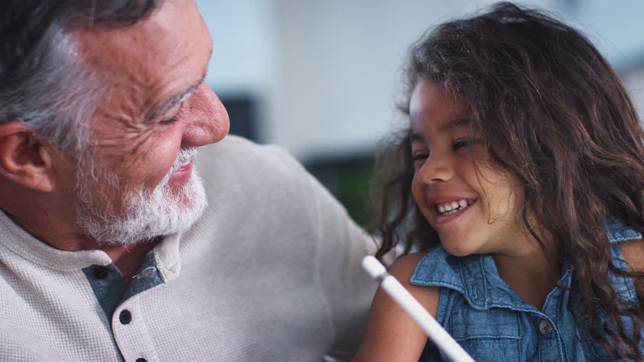 Close up of young girl using tablet computer and stylus, talking with her grandfather beside her