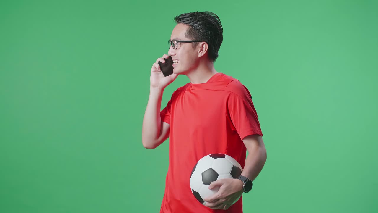 Vista laterale di uomo asiatico con un pallone che parla allo smartphone mentre tifa per il calcio su sfondo green screen