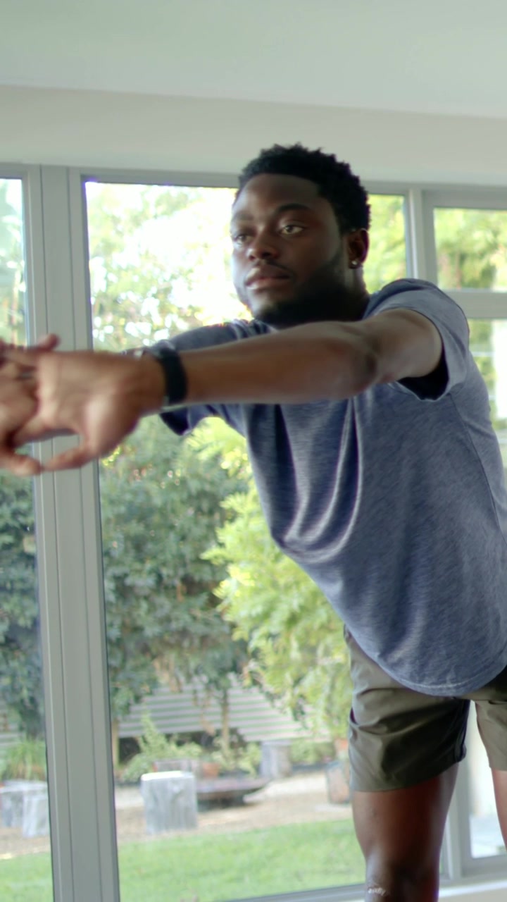Vertical video: Shifting, man in tee and shorts doing single-leg balance at home, watch, window