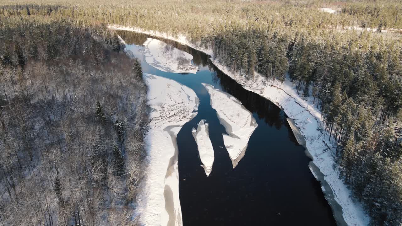 Aerial view of a winding river through a dense snow-covered forest in winter, with a curving road running alongside. A serene and scenic natural landscape.