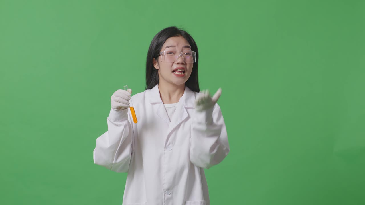 분노한 아시아 여성 과학자가 실험관의 오렌지색 액체를 보고 실험실의 녹색 스크린 배경에 서서 카메라에 소리쳤다.