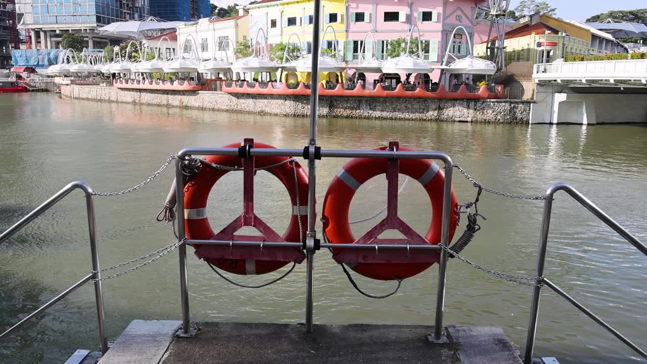 Two red lifebuoys mounted on riverside railings, viewed from concrete steps, with colorful waterfront buildings and calm water in daylight. Static camera