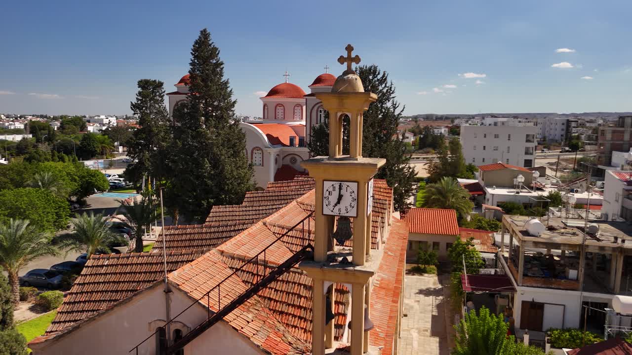 The camera captures a stunning view from above showing a clock tower beside a charming church. The bright blue sky complements the red roofs and lush greenery