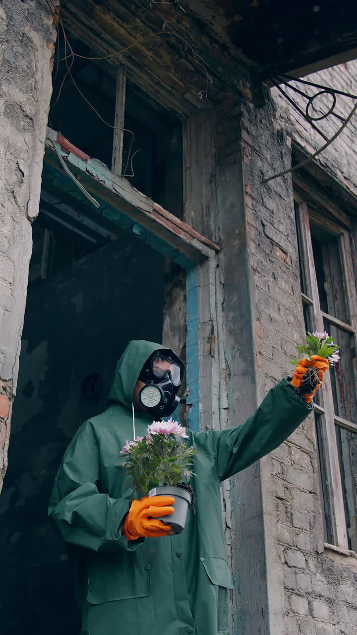 Ecologist standing with plants in abandoned place. Person in protective uniform and gas mask holding flowers near the destroyed building and looking on it. Chemical attack and air pollution concept. Vertical video