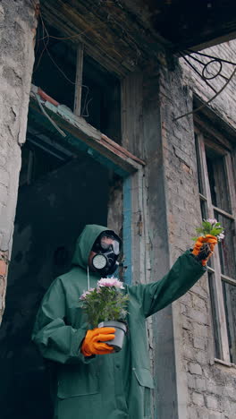 ecologist standing with plants in abandoned place. person in protective uniform and gas mask holding flowers near the destroyed building and looking on it. chemical attack and air pollution concept. vertical video