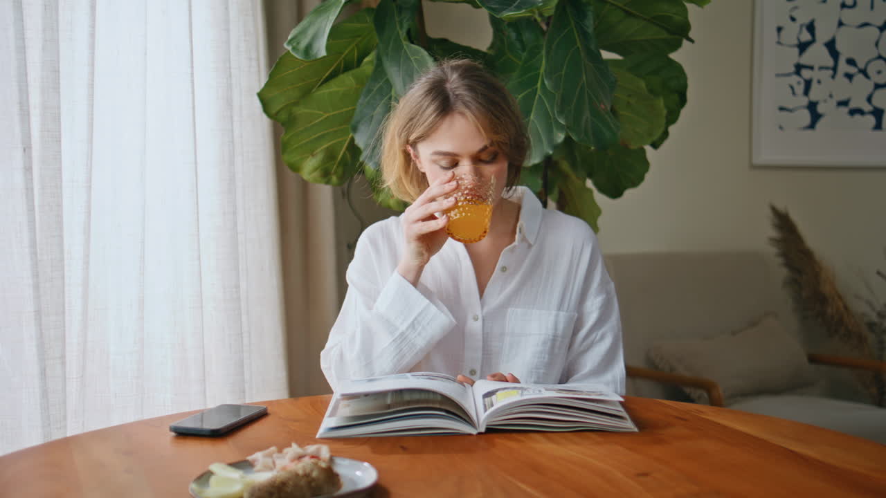 Breakfasting woman reading journal at home kitchen closeup. Lady sipping juice