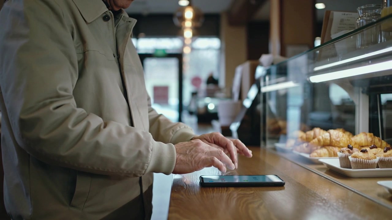 Placing tablet, senior man in beige jacket tapping and typing at cafe counter, with pastry case