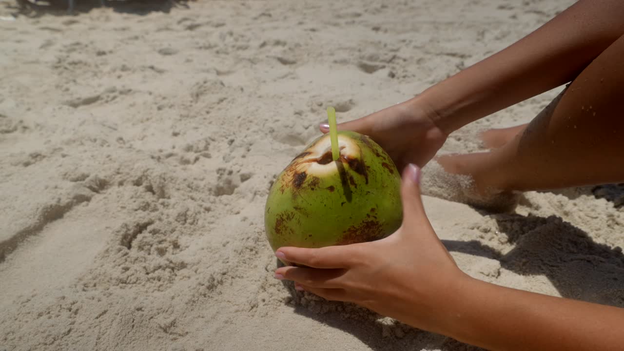 Static closeup of a green coconut sitting on white beach sand. A woman's hands reach into the frame to grab the coconut during early afternoon at Copacabana