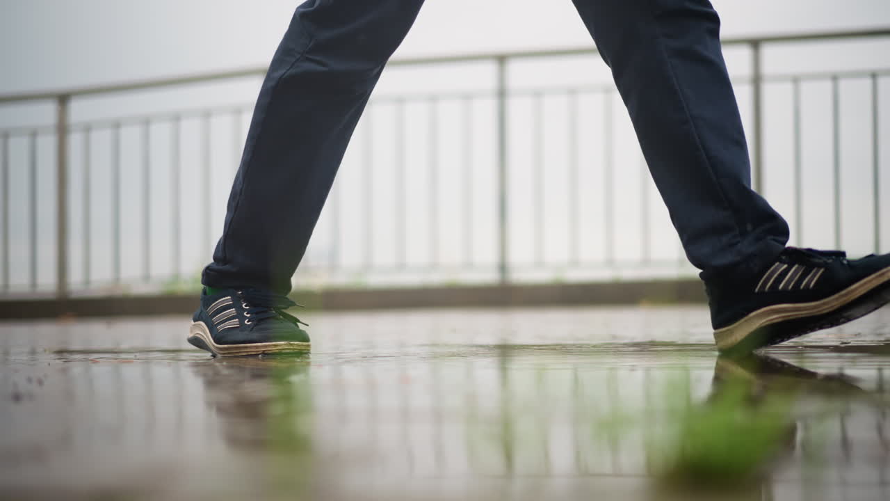 una persona camina por la barandilla de un paseo marítimo mojado; sus pasos firmes crean suaves ondas en los charcos poco profundos; zapatillas y vaqueros empapados en los puños; horizonte apagado bajo nubes cubiertas; ambiente tranquilo y reflexivo