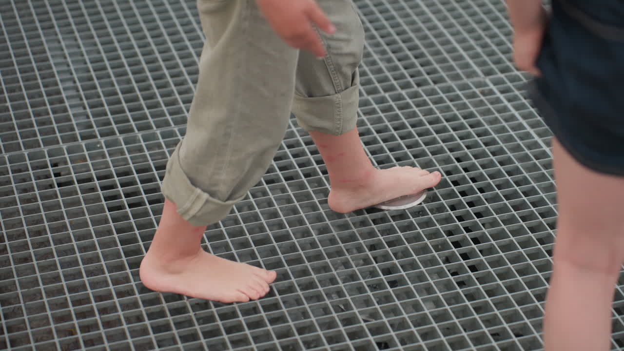Children using feet to close fountain jet on metal grate during picnic, playful leg interaction, water spray responding to pressure, blurred park visitors in background, summer exploration