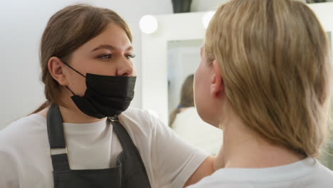 beautician in black mask and white shirt intensely focused on cleaning client brow in well-lit salon. both women maintain eye contact during gentle facial care procedure near illuminated vanity mirror