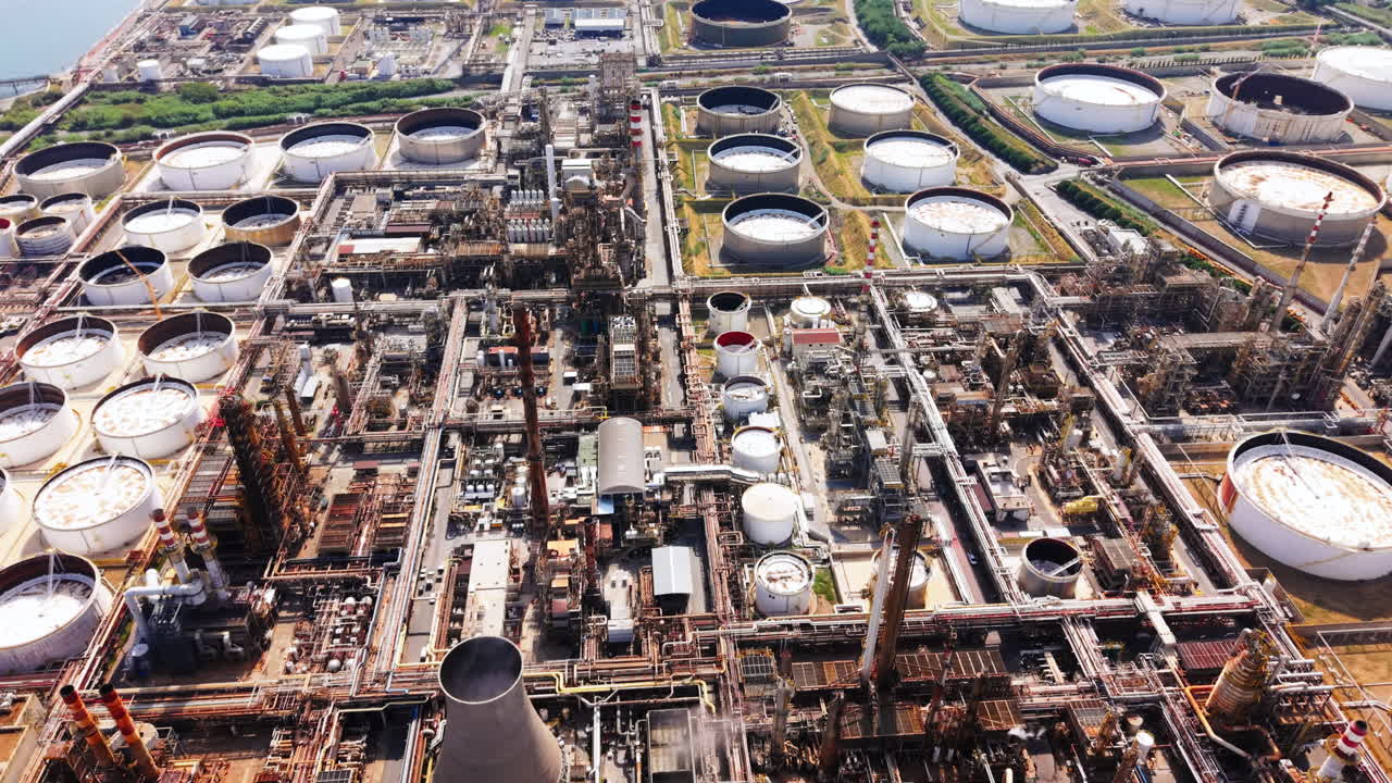 Aerial drone shot in top-down view advancing over a vast oil refinery, showing tanks, pipes, chimneys, and the nearby sea
