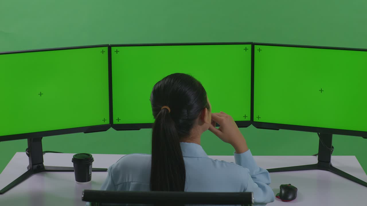 그린 스크린 스튜디오에서 모의 여러 컴퓨터 모니터와 함께 작업하는 동안 무언가에 대해 생각하고 있는 아시아 여성의 뒷면