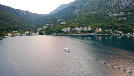 a luxurious white yacht with a tall mast without sails towing an empty motorboat and heading for the port of a small coastal town in the bay of kotor in montenegro, high mountains surrounding the bay