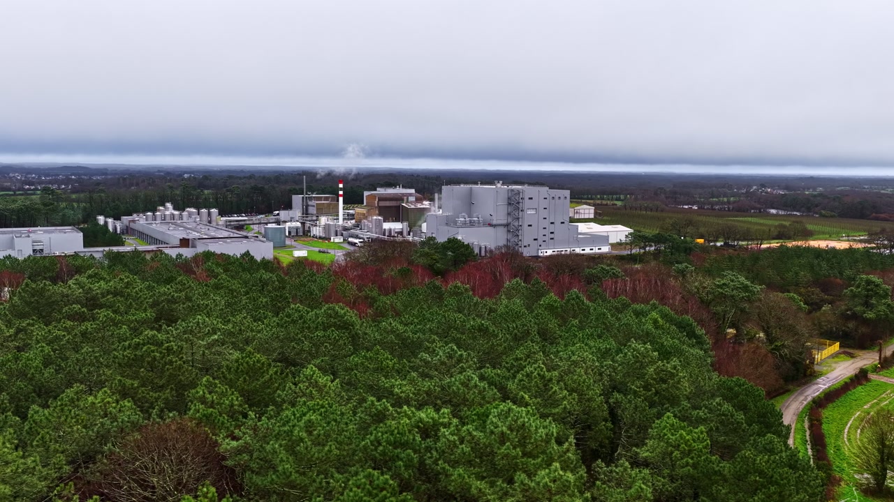 Factory buildings surrounded by trees and farmland, rising above contrasting against forested nature, aerial orbit