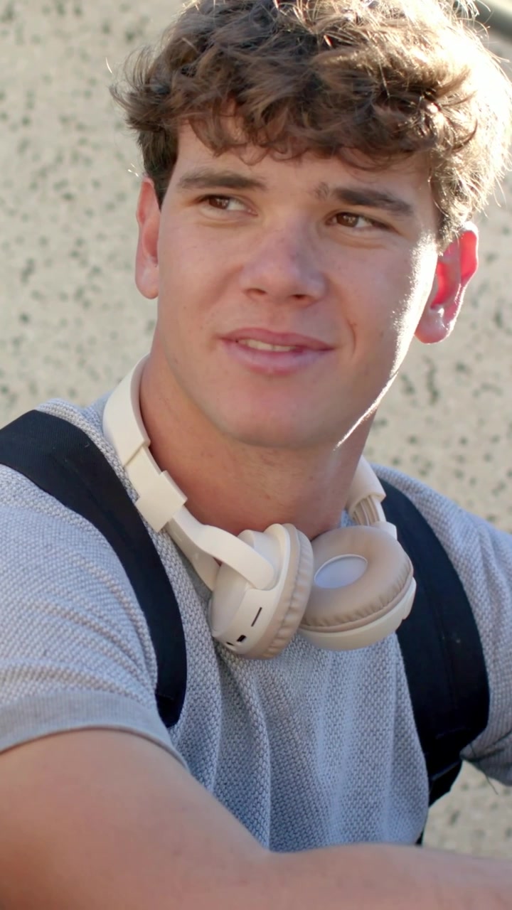 Verticale video: Lachende student in grijs T-shirt met beige headset praat na gebaar van medestudent op de campus.