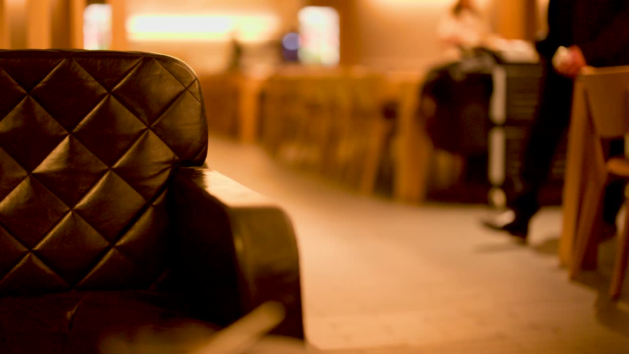 Businessman walks past black leather couch in warmly lit airport lounge, steady camera, shallow focus