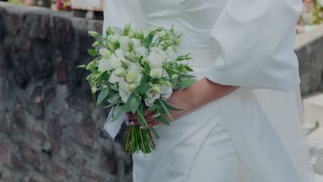 bride holding a bouquet of white flowers and greenery, wearing a white wedding gown, standing near a stone wall