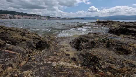 Meereswelle-Spritzt-Auf-Die-Kamerahöhe-über-Den-Felsen-Mit-Blick-Auf-Das-Meer-Und-Die-Stadt-Im-Hintergrund-Hinter-Dem-Meer,-Bewölkter-Tag-Mit-Schwammigen-Wolken,-Aufnahme-Blockiert